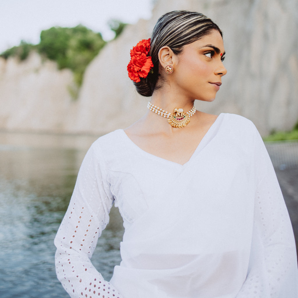 A woman wearing a white chiffon blouse with a floral design, accessorized with a red rose and a golden necklace, posing by a body of water.
